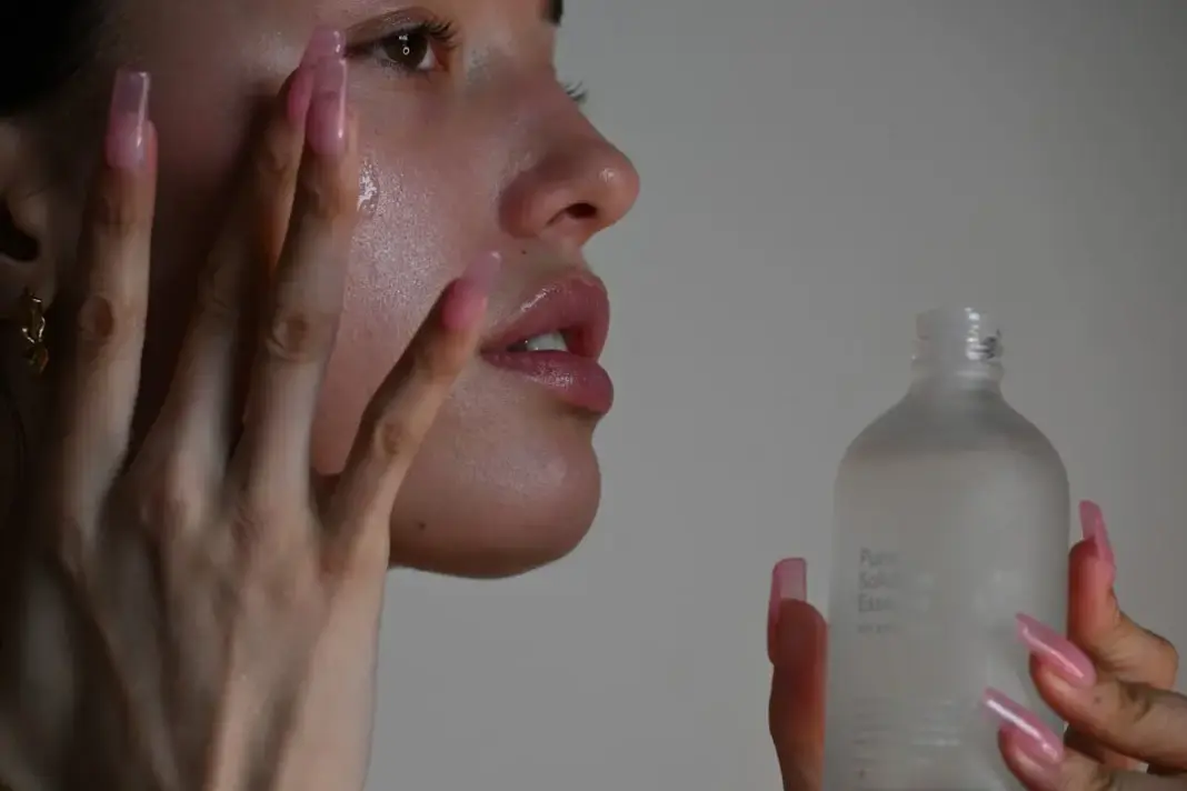 A person applying serum to their cheek with long pink nails